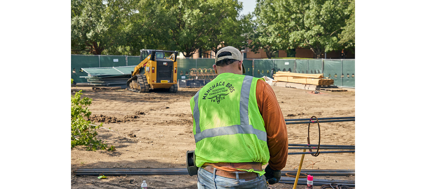 A construction worker wearing a neon safety vest labeled “Menchaca Bros Concrete Construction FWTX” stands at a job site with rebar in front of him and a yellow CAT skid steer in the background.